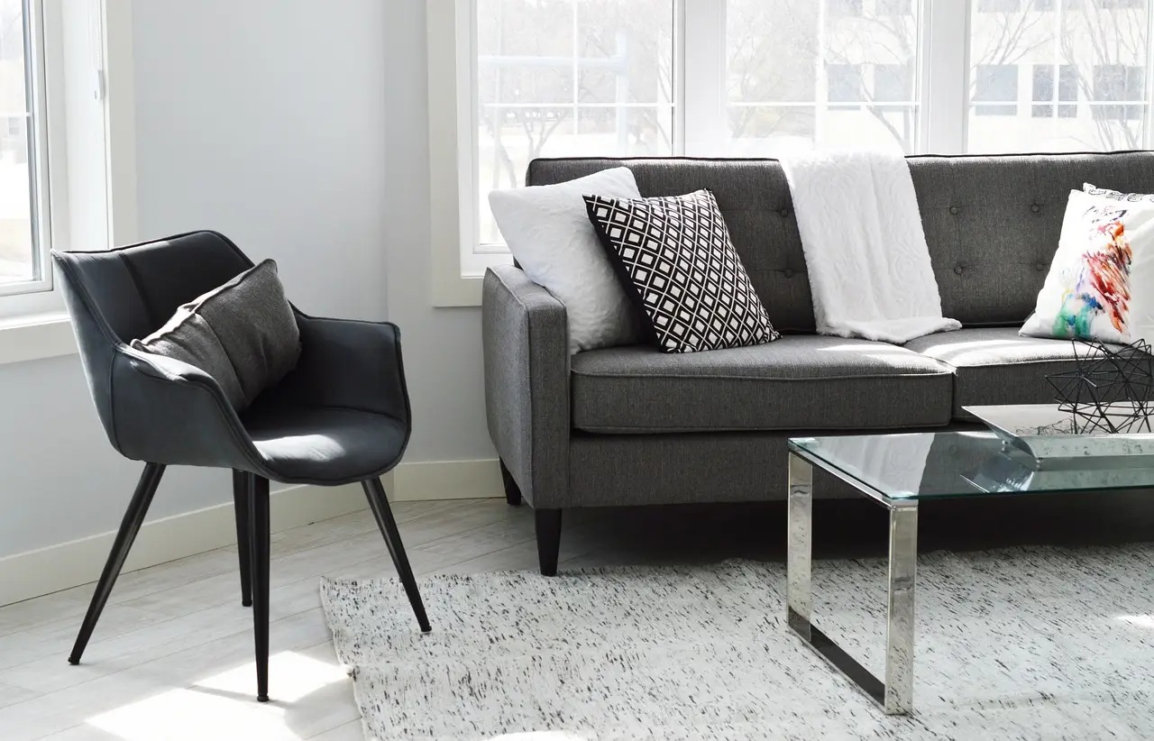 Séjour d'appartement rénové aux murs blancs, canapé gris, fauteuil noir et table basse en verre devant une fenêtre lumineuse